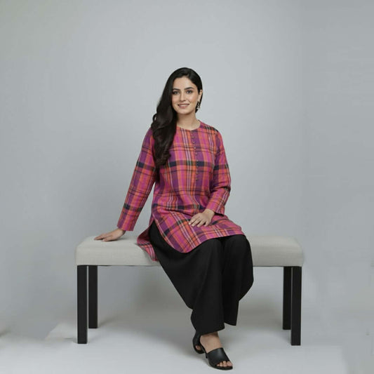 Woman sitting on a bench wearing a pink plaid top and black pants against a gray background